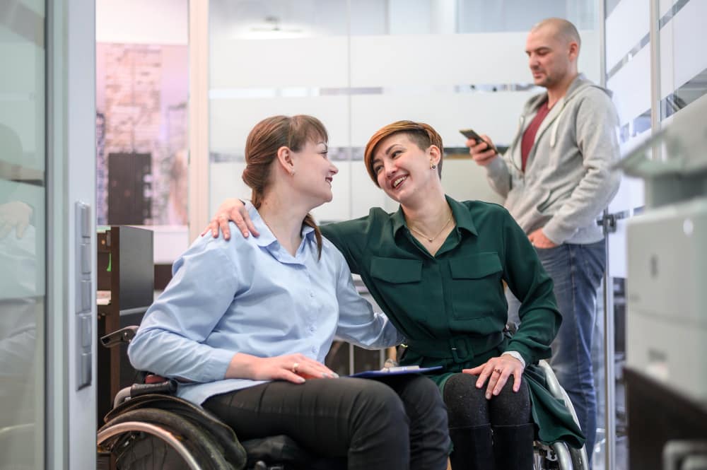 NDIS participants in wheelchairs sharing a supportive moment with a caregiver in an office environment, promoting community participation, social connection, and independent living support.