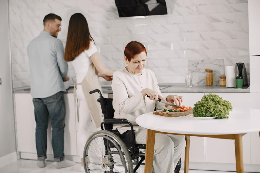 NDIS participant in a wheelchair preparing vegetables in the kitchen with support nearby, building daily living skills and independence through personalised home support.